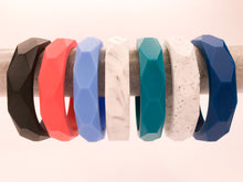 Load image into Gallery viewer, Row of geometric shape teething bangles hanging. Assortment of colours, black bangle, coral bangle, baby blue bangle, marble bangle, teal bangle, denim blue bangle. 
